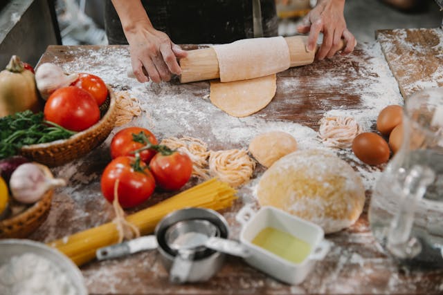 A person's hand making fresh pasta, table is covered with fresh tomatos, flour, eggs and olive oil