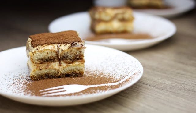 Three individual tiramisu desserts on white plates, featuring layers of coffee-soaked ladyfingers and cream, topped with cocoa powder, with a fork outline drawn in cocoa on the plate in the foreground.