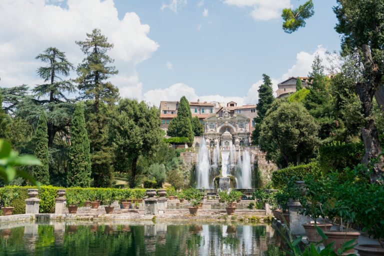 The Fontana dell'Ovato (Oval Fountain) at the Villa d'Este in Tivoli, Italy, showcasing its cascading water features, the terraced gardens with numerous potted plants, and the architectural details of the villa against a backdrop of a partly cloudy sky.