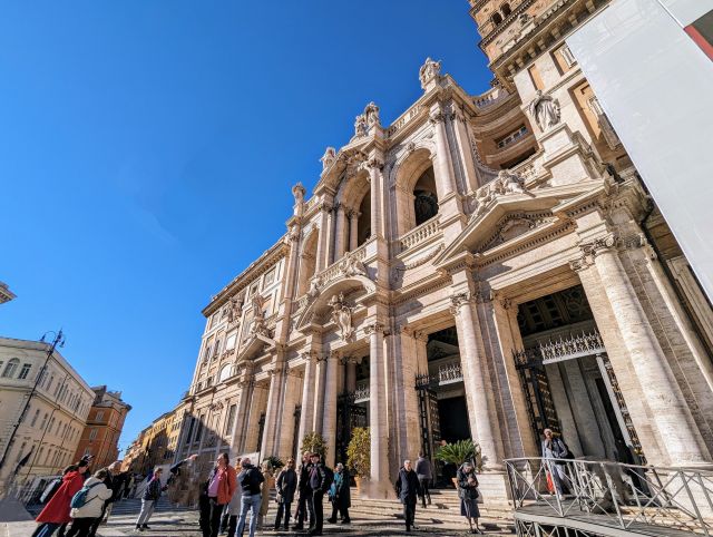The facade of the Papal Basilica of Saint Mary Major (Basilica di Santa Maria Maggiore) in Rome, Italy, featuring a portico with columns, an upper level with windows and an inscription, and a tower rising above, under a clear blue sky.