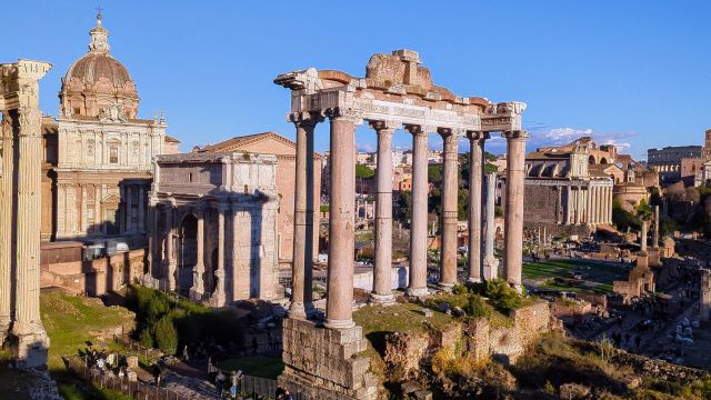 Aerial view of the Roman Forum (Foro Romano) in Rome, Italy, featuring the standing columns of a Roman temple, the ruins of other ancient buildings, and a glimpse of the cityscape in the background, under a clear blue sky.