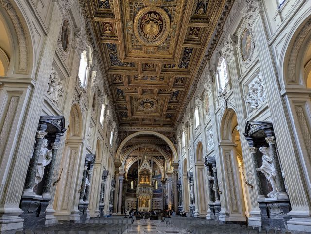 Interior of the Papal Archbasilica of St. John Lateran in Rome, Italy, showcasing the grand central nave with its richly decorated coffered ceiling featuring gilded details, rows of columns separating the side aisles, numerous statues in niches along the nave, and the altar visible in the distance.