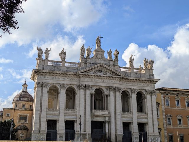The grand facade of the Papal Archbasilica of St. John Lateran in Rome, Italy, featuring towering columns, numerous statues of saints atop the building, the dedicatory inscription and a wide staircase leading up to the entrance.