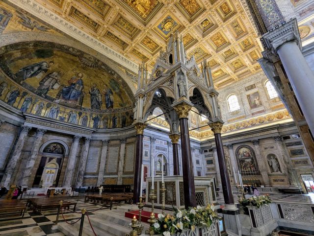 Interior of the Papal Basilica di San Paolo fuori le mura (Basilica of Saint Paul Outside the Walls) in Rome, Italy, showcasing the long central nave with its ornate coffered ceiling, rows of columns separating the side aisles, the prominent mosaic on the wall, the altar area, and other artwork and inscriptions.