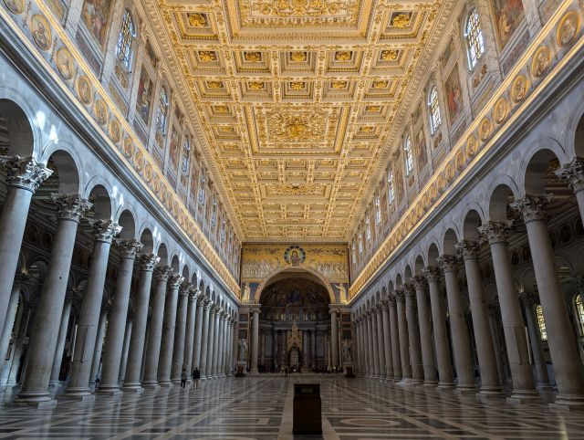 Interior of the Papal Archbasilica of St. John Lateran in Rome, Italy, showcasing the long central nave with its ornate coffered ceiling, rows of columns separating the side aisles, artwork and inscriptions on the walls, and a glimpse of the apse in the distance.