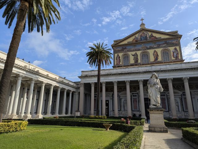 The Basilica di San Paolo fuori le mura (Basilica of Saint Paul Outside the Walls) in Rome, Italy, featuring a large colonnaded courtyard, a church building with a mosaic facade depicting Christ and the Apostles, a statue of Saint Paul in the foreground, and palm trees in the background.