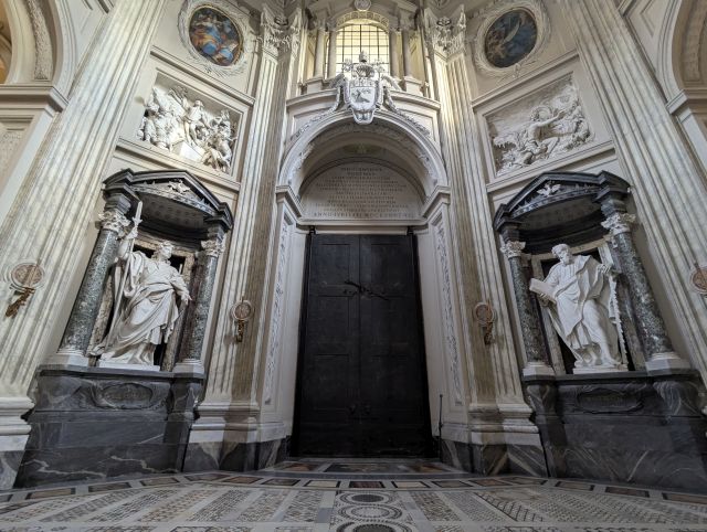 Interior detail of the Papal Archbasilica of St. John Lateran in Rome, Italy, showcasing a large bronze doorway flanked by statues within niches, ornate wall decorations with relief sculptures or paintings, and a patterned marble floor.