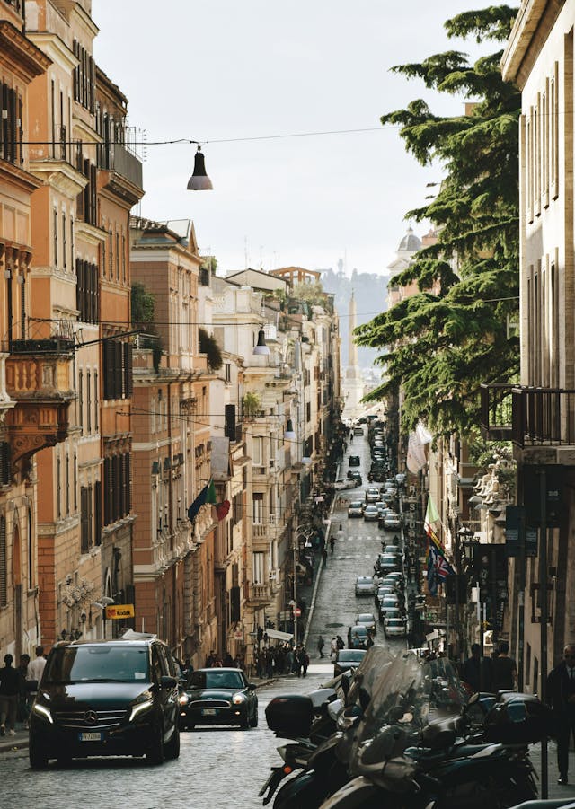 A narrow, cobblestone street in Rome, Italy, lined with tall, multi-story buildings in warm colors, with cars parked along the sides, pedestrians walking, and a glimpse of an obelisk in the distance.