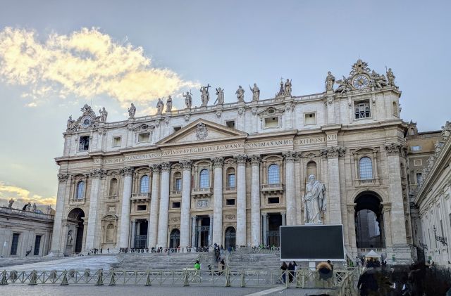 The facade of St. Peter's Basilica in Vatican City, featuring a large central doorway, towering columns, statues of saints atop the building and people at the base of the steps.
