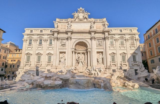 The Trevi Fountain in Rome, Italy, a large Baroque fountain with sculptures of Oceanus and his entourage, cascading water, and architectural details, set against a clear blue sky.
