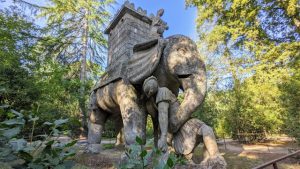 Large, weathered stone sculpture depicting an elephant with a tower-like structure on its back, lifting a figure below.