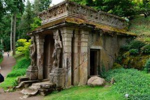 Exterior view of a weathered stone mausoleum with classical architecture and two standing statues at the entrance, surrounded by trees and greenery.
