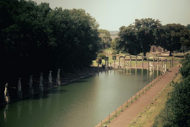 The Canopus at Hadrian's Villa, a UNESCO World Heritage Site in Tivoli, Italy, featuring a long rectangular pool reflecting statues and the ruins of the ancient Roman complex, with trees and hills in the background.
