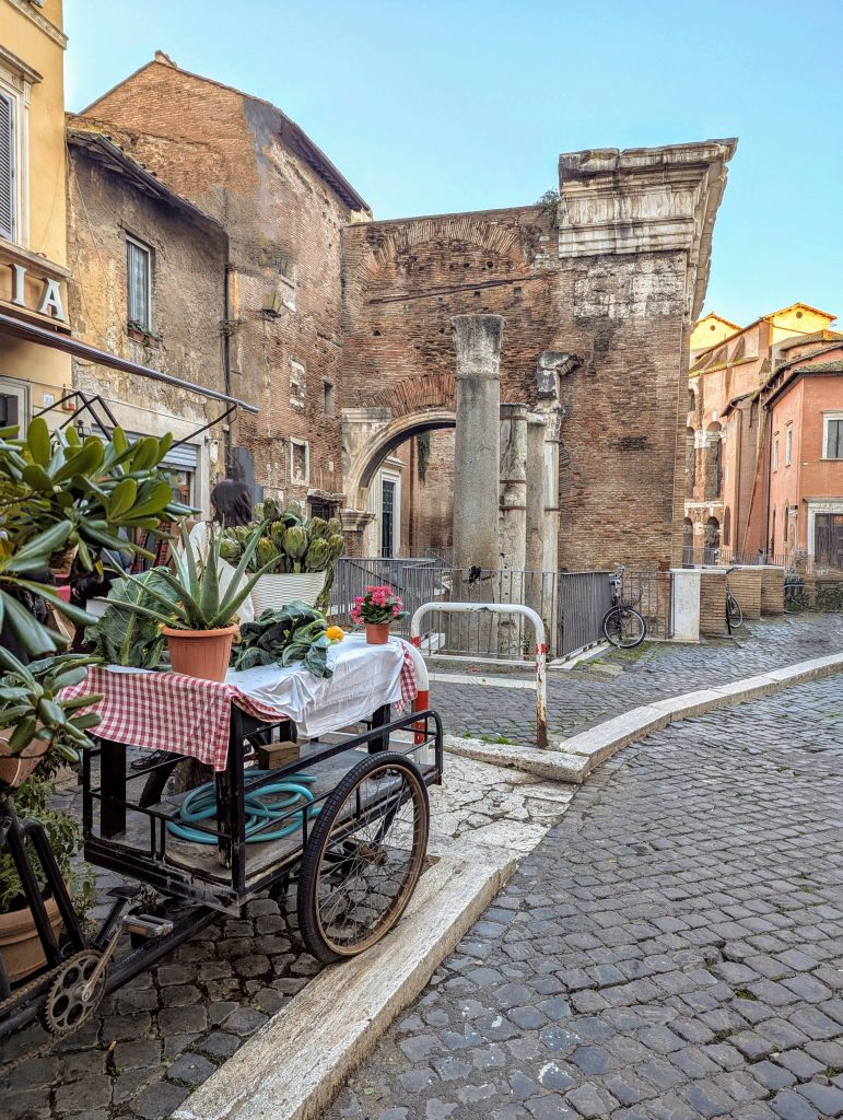 A charming cobblestone street in the Trastevere neighborhood of Rome, Italy, featuring the historic ruins of the Portico d'Ottavia with its arched entrance and remnants of columns, a parked flower cart with colorful blooms, and historic buildings.