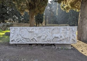 arge, carved Roman sarcophagus featuring a scene of a chariot race or equestrian event, displayed outdoors in Ostia Antica archaeological park with tall trees in the background.