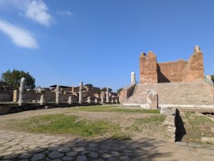 Ruins of Ostia Antica, the ancient port of Rome, in Italy, featuring a prominent brick structure with stairs on the right, and a row of columns from another ruin in the background, under a clear blue sky.