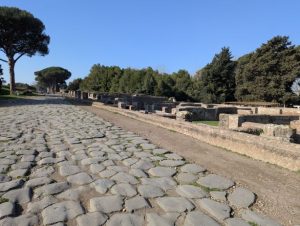 Ancient paved road in Ostia Antica, the ancient port of Rome, Italy, with ruins visible on both sides and characteristic umbrella pine trees in the background, under a clear blue sky.