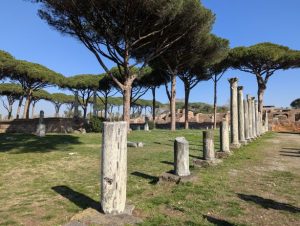 The Piazza delle Corporazioni (Square of the Corporations) in Ostia Antica, the ancient port of Rome, Italy, featuring a series of standing columns bordering an open square with scattered ruins and characteristic umbrella pine trees in the background, under a clear blue sky.