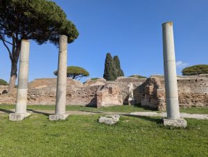 Ruins of Ostia Antica, the ancient port of Rome, in Italy, featuring three prominent columns in the foreground with the remains of brick walls and structures in the background, under a clear blue sky.