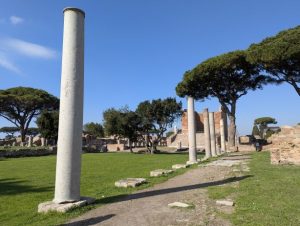 Ruins of Ostia Antica, the ancient port of Rome, in Italy, featuring a prominent column in the foreground, with other columns and the remains of brick walls and structures in the background, along with characteristic umbrella pine trees, under a clear blue sky.