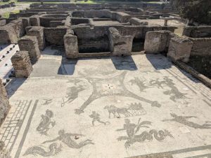 Ruins of the Terme dei Cisiarii (Baths of the Cisiarii) in Ostia Antica, the ancient port of Rome, Italy, featuring a well-preserved black and white mosaic floor with depictions of chariots (cisii) and other figures, surrounded by the remains of brick walls and structures, under a clear blue sky.