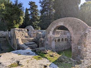 Well-preserved brick ruins with arches and walls at Ostia Antica, the ancient port of Rome, Italy, with lush greenery and trees visible in the background under a blue sky.