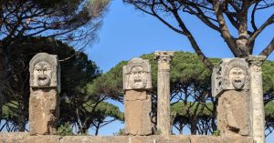 Three ancient Roman theatrical masks, depicting dramatic expressions, at the Teatro di Ostia Antica (Theatre of Ostia Antica) in Italy, with a column and trees visible in the background, under a blue sky.
