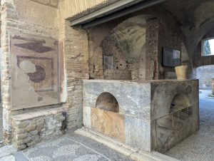 Remains of a thermopolium (ancient Roman food and wine bar) counter) in Ostia Antica, the ancient port of Rome, Italy, featuring a counter with embedded jars (dolia) for food and drinks, constructed of brick and stone, with a partial view of the surrounding ruins.