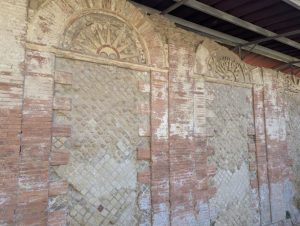Detail of a brick wall with arched openings and decorative elements, showing Roman architectural influences, with visible signs of aging and restoration, under a bright light source.