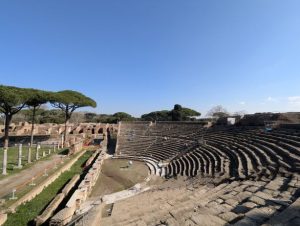 The ancient Roman Theatre of Ostia Antica (Teatro di Ostia Antica) in Italy, showcasing its semi-circular seating area, the stage area, and the surrounding ruins, with characteristic umbrella pine trees visible, under a clear blue sky.